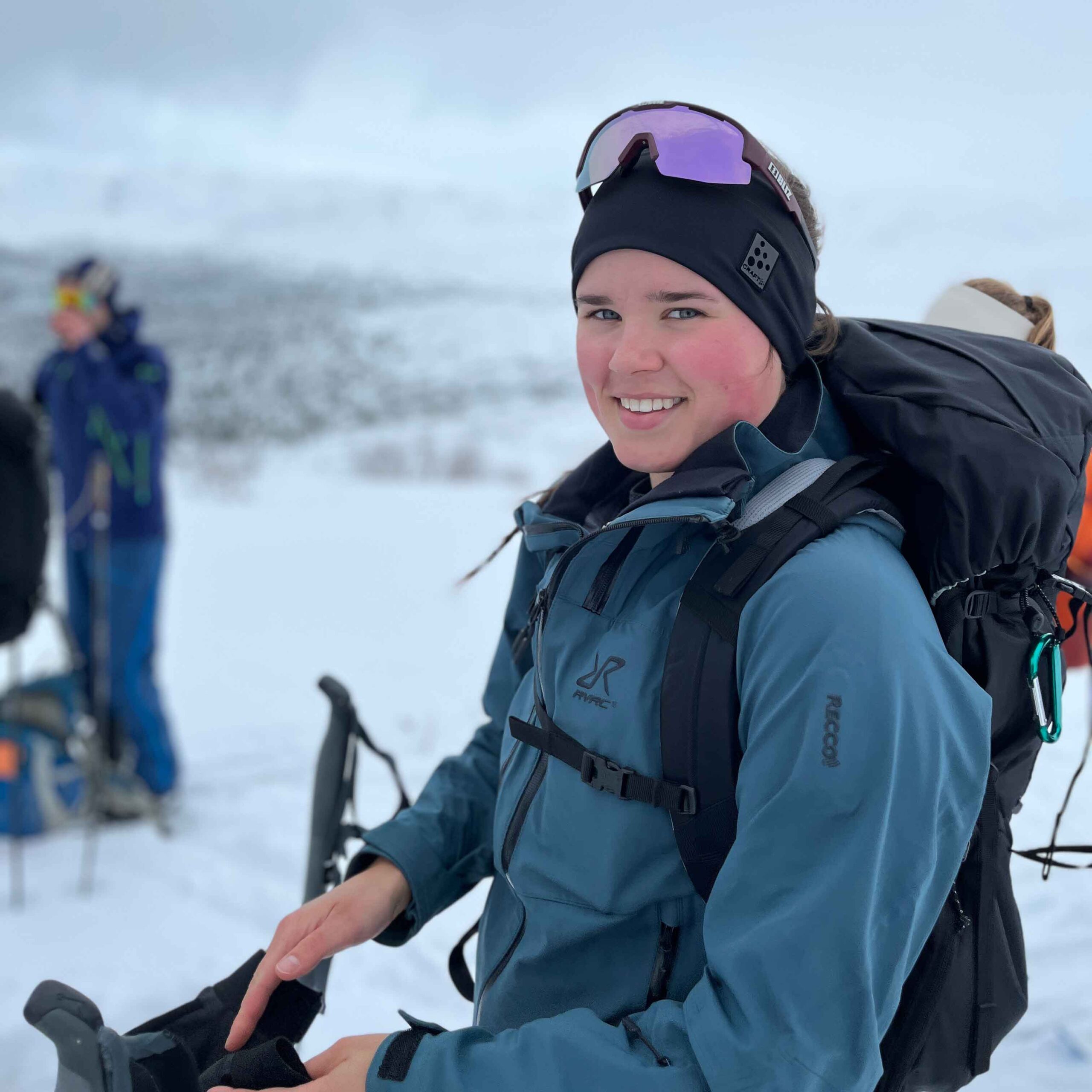 Leende kvinna med ryggsäck och pannband i vinterlandskap, klädd i blå softshell-jacka under en fjälltur